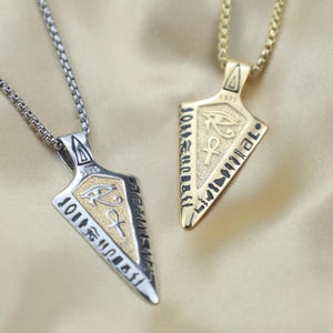 Könnte beinhalten: Zwei Halsketten mit Anhängern im ägyptischen Stil. Eine ist silberfarben, die andere goldfarben. Jede hat einen dreieckigen Anhänger mit Hieroglyphen, dem Horusauge und dem Ankh-Symbol. Die Anhänger hängen an passenden Kettenhalsketten.