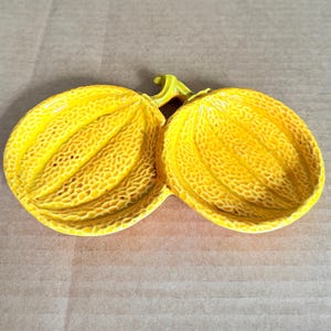 May include: A pair of yellow, melon-shaped decorative objects with textured surfaces and a connecting stem. The objects are a bright yellow color and appear to be made of a solid material. The background is a light brown cardboard.