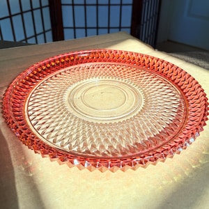 May include: A vintage, clear glass plate with a vibrant red-orange hue. The plate features a diamond-cut pattern in the center and a textured, sawtooth edge. The plate is circular and appears to be a decorative or serving piece.