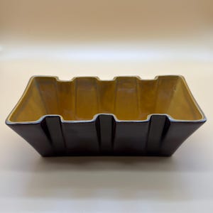 May include: A rectangular ceramic bowl featuring a dark brown exterior and a contrasting mustard yellow interior. The vessel has a distinct geometric design with vertical ridges along the sides and a wide, open top with a scalloped rim edge.