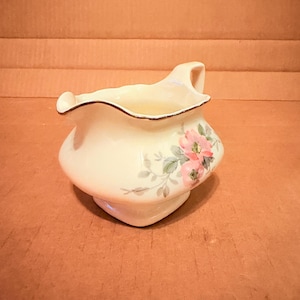 May include: Cream-colored ceramic creamer with a floral design. The creamer has a ruffled rim with a gold trim and a handle. Pink and green floral pattern on the side. The creamer is a decorative piece for serving cream or milk.