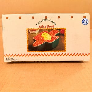 May include: A white cardboard box with the words "Terra Cotta Cactus Salsa Bowl" printed on it. The box features an image of a green cactus-shaped bowl filled with salsa and a dollop of sour cream. The box has a brown wooden background.