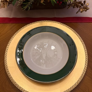 May include: A gold charger plate with a rope-like edge, topped with a green and gold rimmed plate, and a white bowl. The arrangement is set on a wooden table, with a festive floral arrangement in the background.