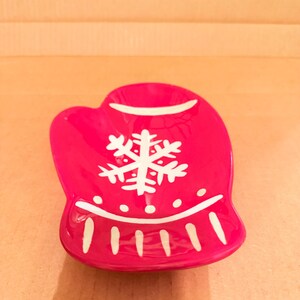 May include: A red ceramic mitten-shaped dish with a white snowflake design and white trim. The dish is a festive holiday decoration, perfect for serving small treats or holding jewelry.