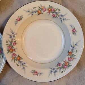 May include: Off-white ceramic soup bowls with a floral design. The bowls have a gold rim and are decorated with pink, yellow, and blue flowers. The floral pattern is arranged around the rim of the bowl.