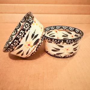 May include: Two ceramic bowls with a cream-colored base and a floral design. The bowls have a black and brown patterned rim and are decorated with black leaves and small yellow and red dots. The bowls are likely used for serving food.