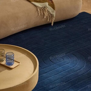 Op de afbeelding: Een moderne woonkamerscène met een marineblauw tapijt met een abstract ontwerp. Een beige bank en een ronde, lichtgekleurde salontafel met een dienblad en glazen zijn ook zichtbaar. Een gestreepte plaid met franjes ligt op de bank.