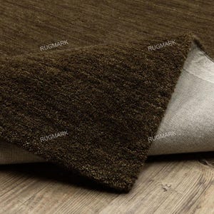 Op de afbeelding: Close-up van een bruin tapijt met een getextureerd oppervlak. Het tapijt is omgevouwen en onthult een lichtere achterkant. Het woord "RUGMARK" staat op verschillende plaatsen op het tapijt gedrukt. Het tapijt ligt op een houten ondergrond.