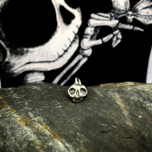 May include: A small silver skull charm pendant, with a detailed design, sits on a dark gray rock.