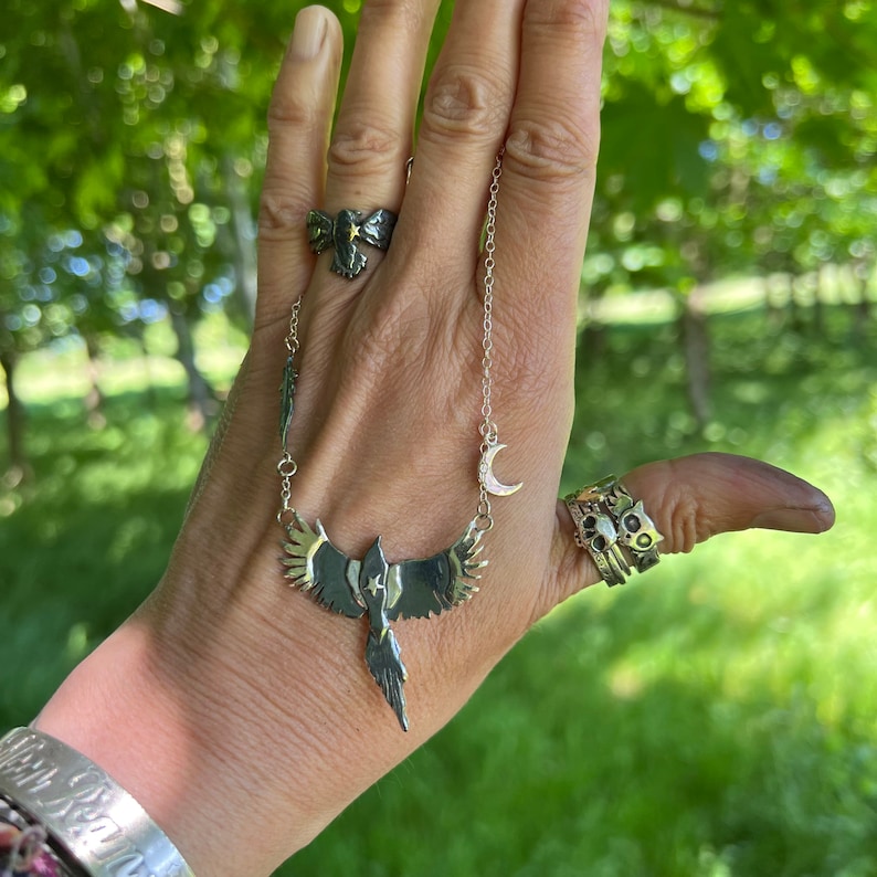 K&ouml;nnte beinhalten: Eine silberne Halskette mit einem Vogelanh&auml;nger, einem Mondsichel-Anh&auml;nger und zwei Ringen. Der Vogelanh&auml;nger hat goldfarbene Fl&uuml;gel. Ein Ring hat ein Schleifendesign mit einem goldenen Stern. Der andere Ring hat ein Katzen-Design.