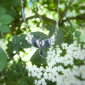 K&ouml;nnte beinhalten: Silberfarbene Halskette mit einem stilisierten Ph&ouml;nix-Anh&auml;nger mit ausgebreiteten Fl&uuml;geln und einem Stern. Die Halskette hat eine Kette und einen Federanh&auml;nger. Der Schmuck ist vor gr&uuml;nem Laub und wei&szlig;en Bl&uuml;ten platziert.