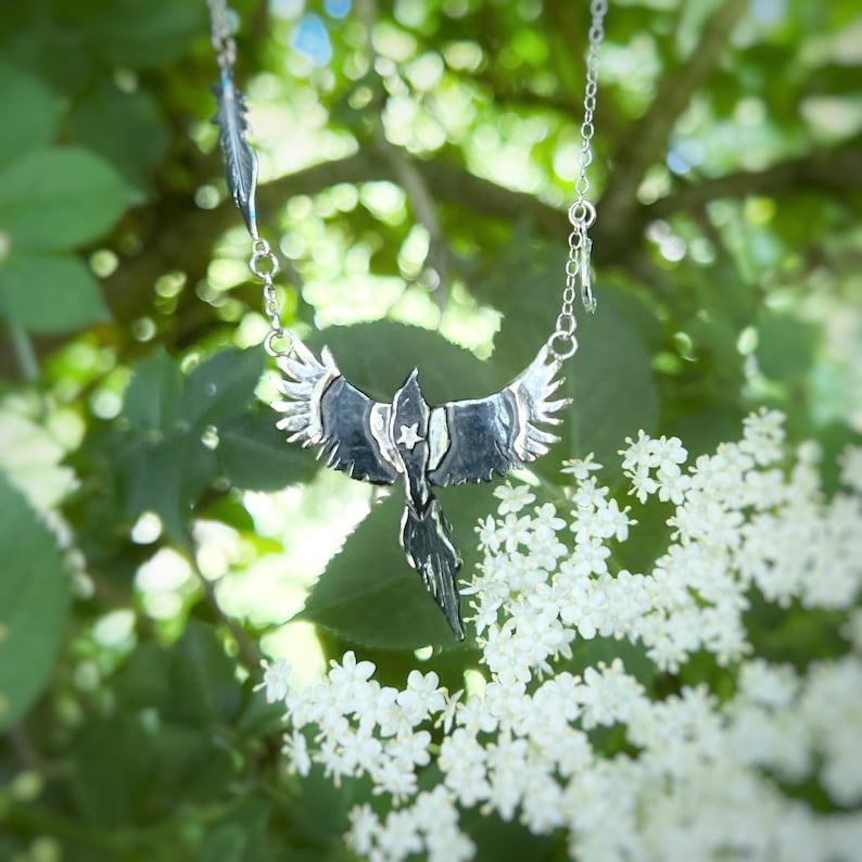 K&ouml;nnte beinhalten: Silberfarbene Halskette mit einem stilisierten Ph&ouml;nix-Anh&auml;nger mit ausgebreiteten Fl&uuml;geln und einem Stern. Die Halskette hat eine Kette und einen Federanh&auml;nger. Der Schmuck ist vor gr&uuml;nem Laub und wei&szlig;en Bl&uuml;ten platziert.