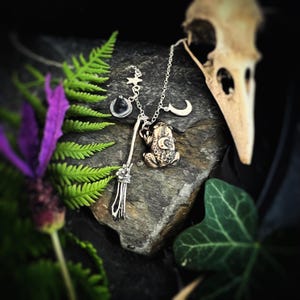 May include: A silver-toned metal necklace featuring a frog pendant with a crescent moon detail, a small witch hat charm, a star charm, a crescent moon charm, and a broomstick charm, all suspended from a delicate silver-toned metal chain.
