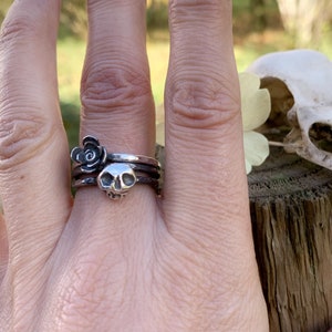 May include: A silver skull ring with a black rose charm on a finger. The ring is worn on the middle finger of a hand.