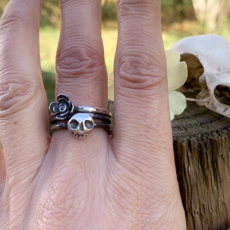 May include: A silver skull ring with a black rose charm on a finger. The ring is worn on the middle finger of a hand.