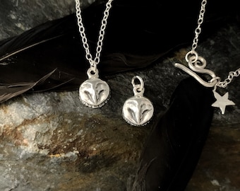 May include: Two silver owl pendant necklaces and a single owl pendant. The pendants are round with a detailed owl face design. One necklace has a chain with a star charm. The background includes a black feather and a dark stone surface.