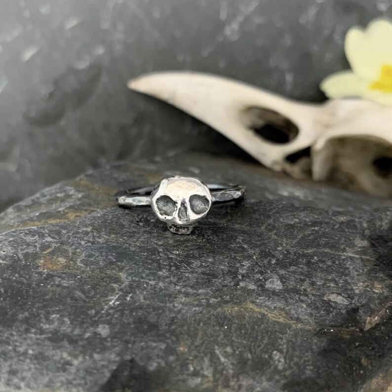 May include: A silver skull ring with a textured finish. The ring is on a gray rock.