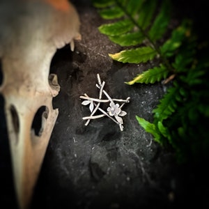 May include: A silver five-pointed star pendant with a small flower in the center. The star is made of twisted wire and has a rustic, organic look.