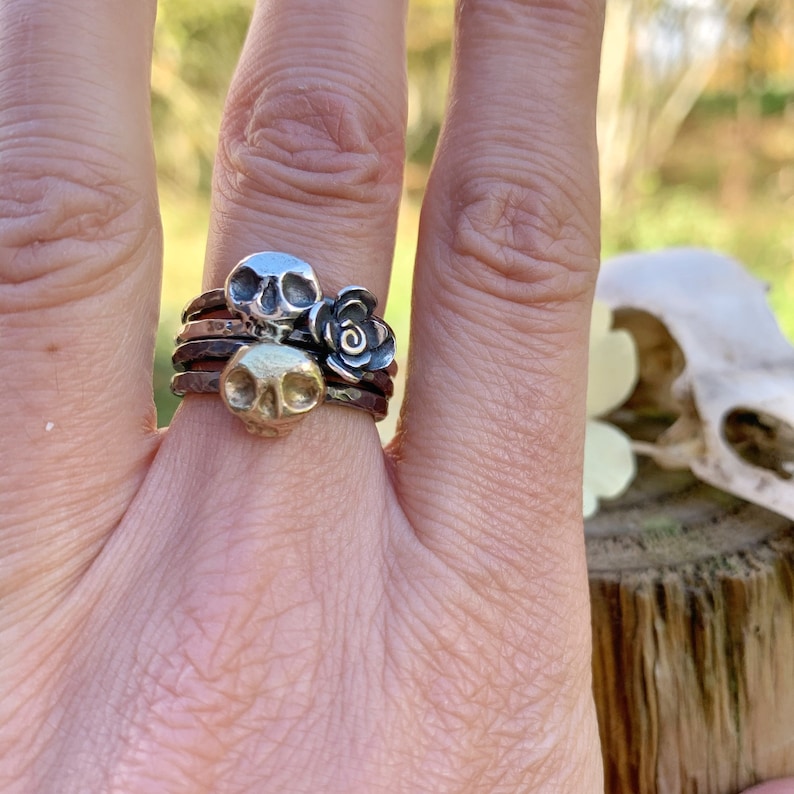 May include: A silver ring with three stacked bands, featuring two small skull charms and a rose charm.