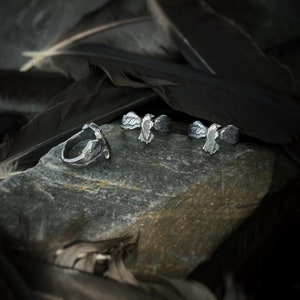 May include: Four silver rings featuring a bird design. The rings are arranged in a row on a grey rock. The rings are made of metal and have a detailed design.