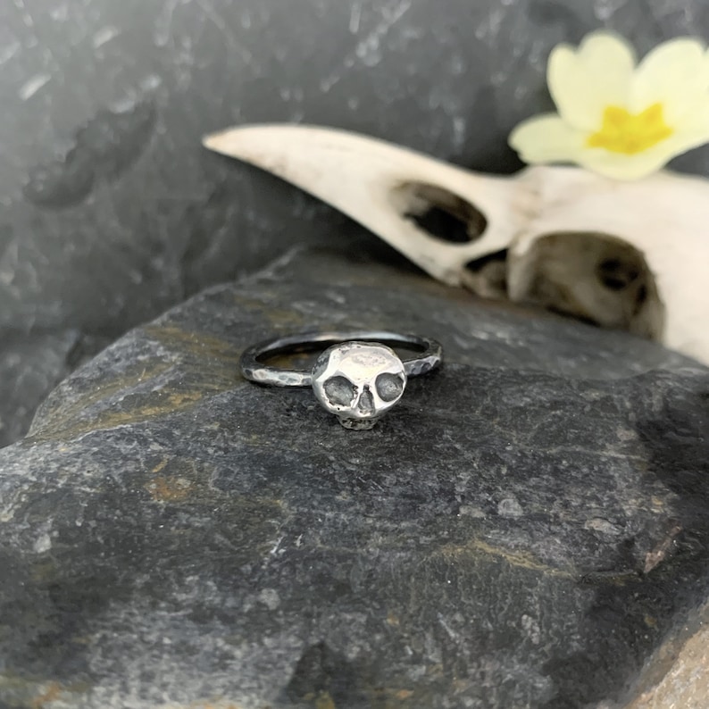 May include: A silver skull ring with a textured finish. The ring is on a dark gray rock.