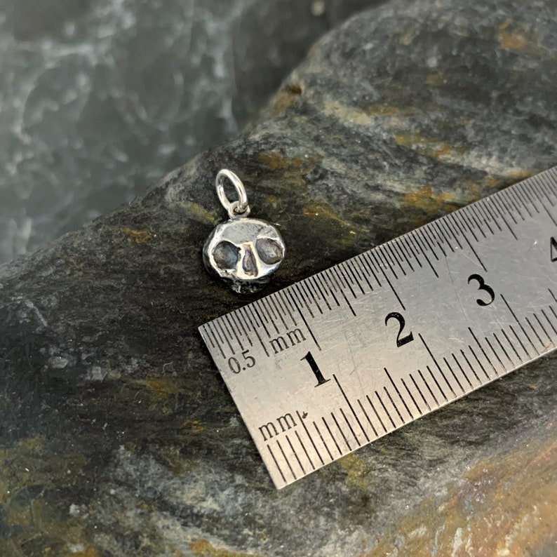 May include: A silver skull charm pendant measuring approximately 1 cm in diameter. The charm is shown next to a ruler for size reference.