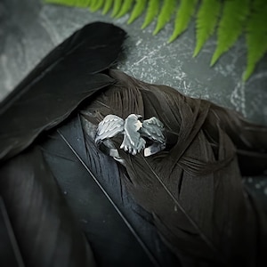 May include: A silver ring featuring a detailed design of a raven with its wings outstretched. The ring is displayed against a backdrop of black feathers.