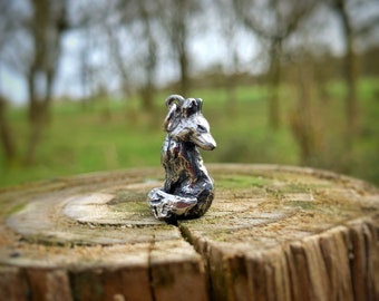 May include: A silver fox charm, sitting on a wooden stump, with a blurred background of trees.
