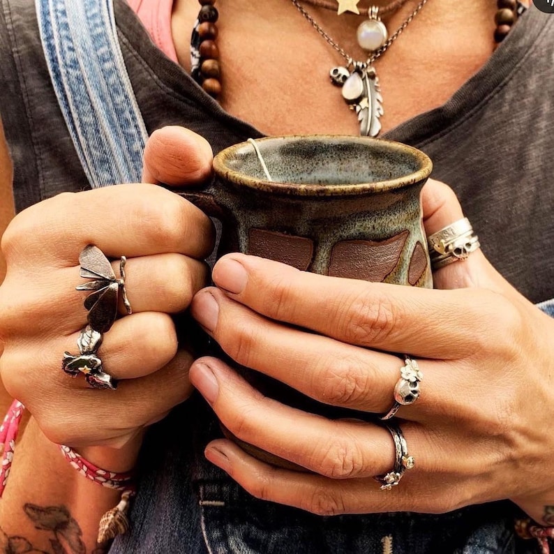 May include: A person's hands holding a brown ceramic mug with a tea bag. The person is wearing several silver rings with butterfly, flower, and skull designs.