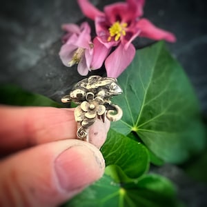 May include: A silver ring with a frog design, set with a pink gemstone and floral details. The ring is held up, with pink flowers and green leaves in the background.