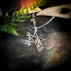 May include: A silver pendant necklace featuring a star and floral design. The pendant has a star at the top, a floral element, and a geometric design. The necklace is displayed on a dark, textured surface with green foliage.