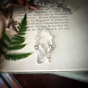 May include: A silver charm featuring two rabbits, a bird, and stars. The charm is in the shape of a crescent moon and is hanging from a silver chain.