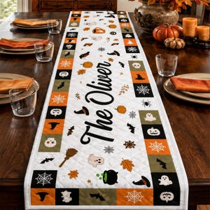 Pode incluir: Um caminho de mesa branco acolchoado com o texto The Oliver em letra preta, rodeado por um padrão de quadrados laranja, pretos e verde-azeitona com ícones de Halloween como fantasmas, abóboras, teias de aranha, caveiras e chapéus.