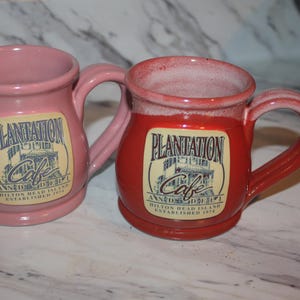 May include: Two ceramic mugs, one pink and one red, each featuring a cream-colored label with the text Plantation Cafe, Ann Dehi, Hilton Head Island, Established 1974. Both mugs have a rounded body, a wide rim, and a curved handle on the side.