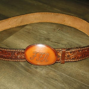 May include: A brown leather belt featuring an oval-shaped buckle with the engraved letters FFN. The belt strap displays a detailed floral tooling pattern along its length, with visible stitching along the edges and a smooth, tan interior lining.