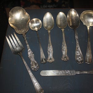 May include: A collection of seven silver-toned metal utensils including a large serving fork, a wide serving spoon, a small ladle, three various sized spoons, and a flat-bladed knife. Each piece features ornate embossed floral handle detailing.