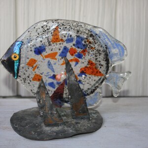May include: A decorative glass fish sculpture. The fish has a clear body with orange, blue, and white accents. It is mounted on a dark gray metal base. The fish's eye is yellow and black.