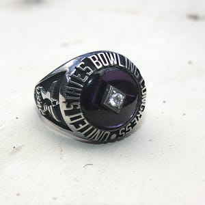 May include: A silver-toned ring with the text "UNITED STATES BOWLING CONGRESS" encircling a dark purple center stone. A small, square, clear stone is set in the center of the purple stone. The ring has detailed side designs.