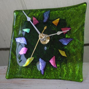 May include: A square, green glass clock with colorful, abstract accents. The clock face features a gold center and white hands. The decorative clock is approximately 5 inches wide.