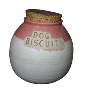 May include: A round ceramic jar with a cork lid, featuring a two-tone design with a dusty pink upper section and a white lower section. The text DOG BISCUITS is embossed on a rectangular plaque on the front of the jar, which has a smooth finish.