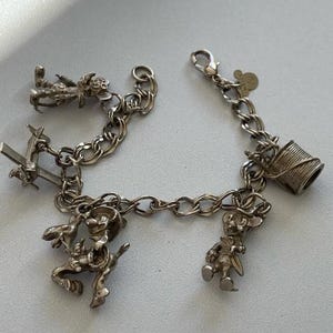Puede incluir: Una pulsera de plata con varios dijes, incluyendo un vaquero a caballo, una bobina de hilo, un avi&oacute;n peque&ntilde;o y un personaje con sombrero. La pulsera tiene un dise&ntilde;o de cadena y un cierre.