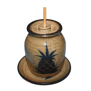 May include: A ceramic butter dish with a lid and a small wooden butter knife. The dish is tan with a dark blue pineapple design. The dish sits on a matching saucer. The lid has a hole for the knife.