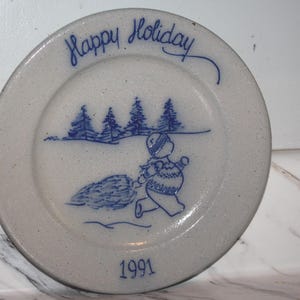 May include: A round, speckled off-white ceramic plate featuring blue hand-painted details. The text "Happy Holiday" is at the top, with a figure carrying a pine tree in a snowy forest scene in the center and the year "1991" printed at the bottom.