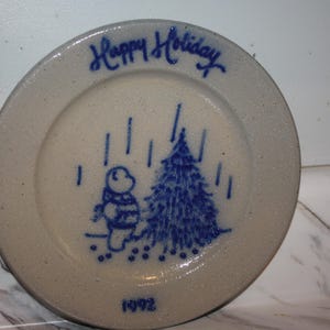 May include: A round, speckled beige ceramic plate featuring blue hand-painted details. The design includes a bear standing next to a Christmas tree under falling snow, the text "Happy Holiday" at the top, and the year "1992" at the bottom.