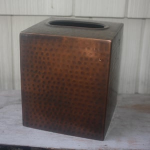 May include: A square tissue box cover featuring a dark, metallic copper finish with a distinct hammered texture across all visible sides. The top surface includes a centered, elongated oval opening designed for dispensing tissues from the container.