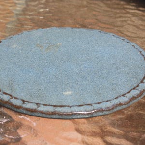 May include: A circular ceramic coaster with a textured, matte blue surface and a dark brown, scalloped edge detail. The coaster features a speckled finish across the center, providing a consistent, flat profile with a decorative border design.