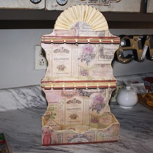 May include: A decorative wooden shelf with a vintage design. The shelf features a fan-shaped top and is adorned with floral and label-style graphics in shades of pink, beige, and brown. It has multiple levels for display and a small bottom shelf.