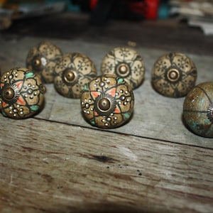 May include: A collection of decorative cabinet knobs with a vintage aesthetic. The knobs feature a round shape with intricate floral and geometric patterns in shades of beige, brown, and teal. Each knob has a brass-colored center and is arranged on a wooden surface.