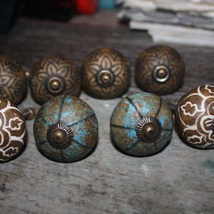 May include: A collection of eight decorative cabinet knobs. The knobs feature a variety of designs, including floral and geometric patterns. Colors include bronze, white, and turquoise. Each knob has a brass-colored base.
