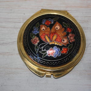 May include: A gold-toned compact mirror with a black enamel top. The top features a butterfly design in orange, red, and yellow, surrounded by colorful flowers. The compact is round and has a hinged opening.